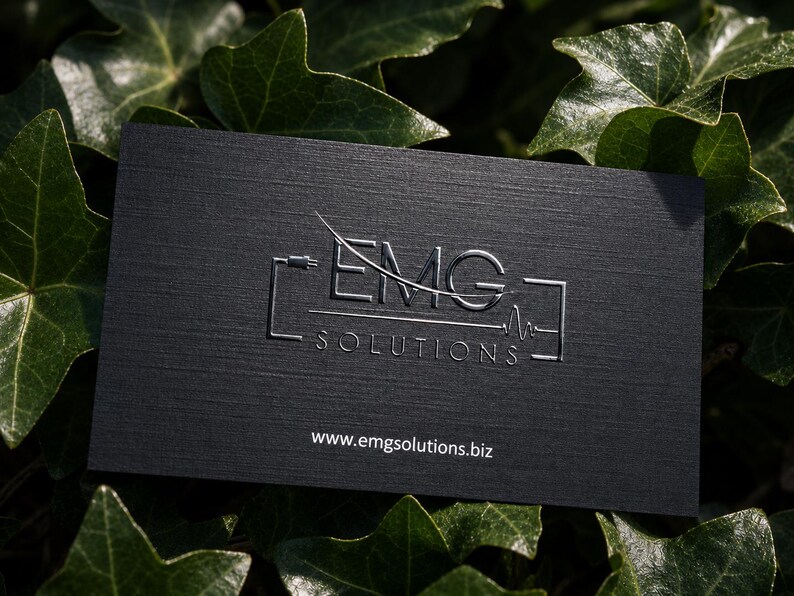 K&ouml;nnte beinhalten: Eine rechteckige schwarze Visitenkarte mit strukturierter Oberfl&auml;che und einem silbernen Pr&auml;gelogo mit der Aufschrift EMG SOLUTIONS sowie einer stilisierten Grafik aus Netzstecker und Pulslinie. Die Website www.emgsolutions.biz ist unten wei&szlig; aufgedruckt.