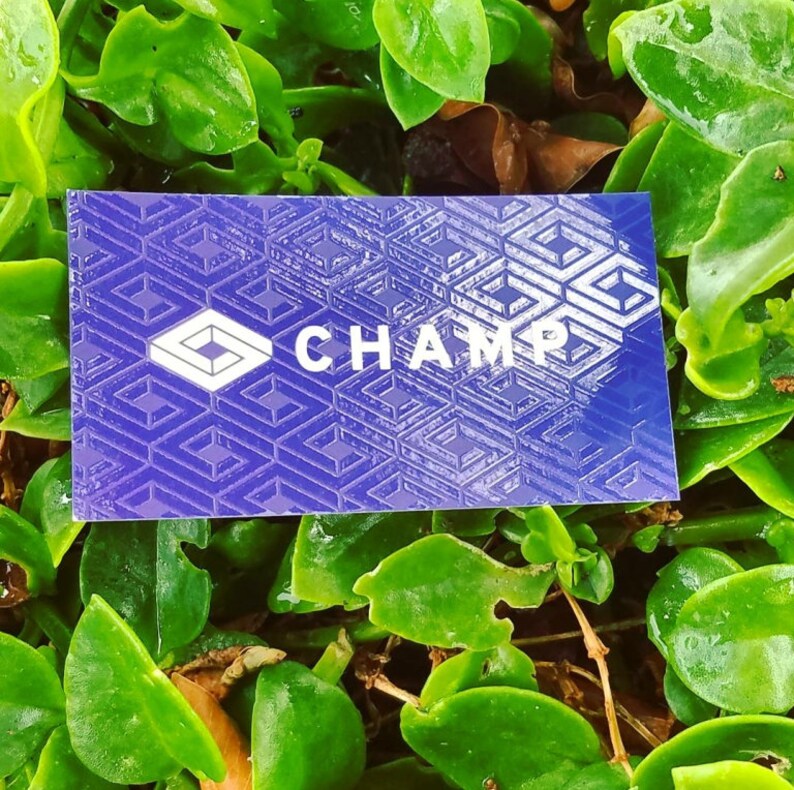 May include: A rectangular business card with a deep purple background featuring a repeating geometric pattern of white interlocking squares. The word CHAMP is printed in white capital letters next to a white diamond-shaped logo icon.