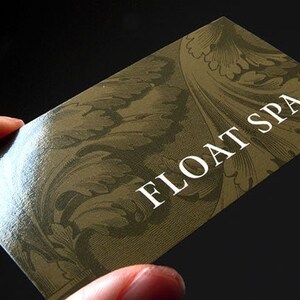 May include: A rectangular business card with a glossy finish, featuring a dark olive green background with a decorative, intricate leaf pattern in a lighter shade of green. The text FLOAT SPA is printed in a bold, white, sans-serif font.