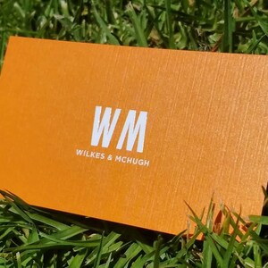 K&ouml;nnte beinhalten: Eine rechteckige orangefarbene Visitenkarte mit strukturierter Oberfl&auml;che. Die Karte zeigt die wei&szlig;en Buchstaben WM in einer gro&szlig;en Schriftart, mit dem Text WILKES & MCHUGH in einer kleineren wei&szlig;en Schrift direkt darunter.