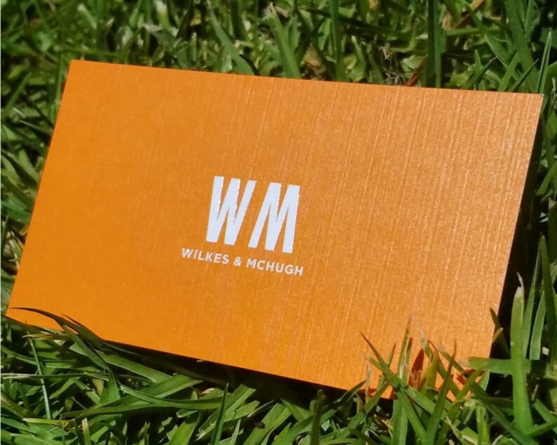 K&ouml;nnte beinhalten: Eine rechteckige orangefarbene Visitenkarte mit strukturierter Oberfl&auml;che. Die Karte zeigt die wei&szlig;en Buchstaben WM in einer gro&szlig;en Schriftart, mit dem Text WILKES & MCHUGH in einer kleineren wei&szlig;en Schrift direkt darunter.