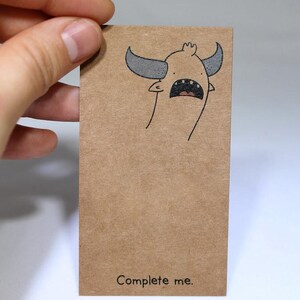 May include: A rectangular brown kraft paper card featuring a black ink illustration of a horned creature with an open mouth at the top and the text "Complete me." printed in black at the bottom centre of the card surface.