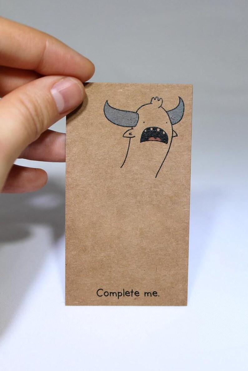 May include: A rectangular brown kraft paper card featuring a black ink illustration of a horned creature with an open mouth at the top and the text "Complete me." printed in black at the bottom centre of the card surface.