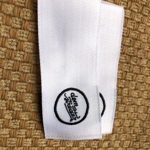 May include: Two rectangular white fabric clothing labels featuring a black embroidered circular logo. Inside the circle, the text "dem theater" is written in a stylized, cursive black font. The labels have a woven texture and clean edges.