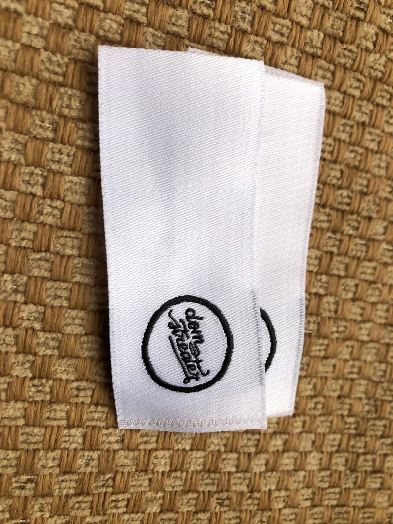May include: Two rectangular white fabric clothing labels featuring a black embroidered circular logo. Inside the circle, the text "dem theater" is written in a stylized, cursive black font. The labels have a woven texture and clean edges.