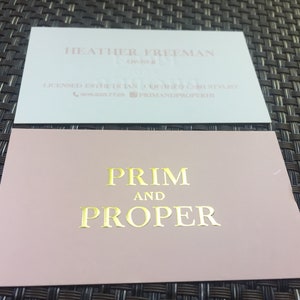 Puede incluir: Dos tarjetas de visita rectangulares. La tarjeta superior es blanca con texto claro que dice HEATHER FREEMAN, OWNER, LICENSED ESTHETICIAN, CERTIFIED LASH STYLIST, un icono de tel&eacute;fono con 508.228.7728 y un icono de Instagram con PRIMANDPROPERHI. La tarjeta inferior es rosa empolvado con texto en l&aacute;mina dorada que dice PRIM AND PROPER.