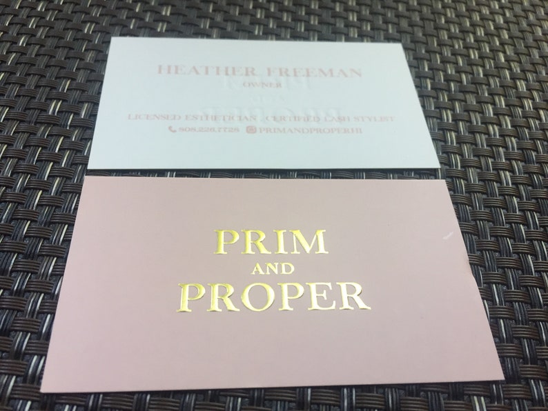 Puede incluir: Dos tarjetas de visita rectangulares. La tarjeta superior es blanca con texto claro que dice HEATHER FREEMAN, OWNER, LICENSED ESTHETICIAN, CERTIFIED LASH STYLIST, un icono de tel&eacute;fono con 508.228.7728 y un icono de Instagram con PRIMANDPROPERHI. La tarjeta inferior es rosa empolvado con texto en l&aacute;mina dorada que dice PRIM AND PROPER.