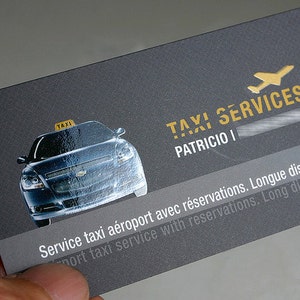 May include: A rectangular business card with a dark gray textured background. It features a glossy graphic of a black taxi with a yellow roof sign. The text reads TAXI SERVICES, PATRICIO, and Service taxi a&eacute;roport avec r&eacute;servations.