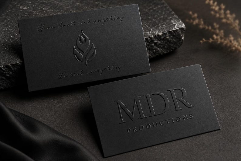 May include: Two rectangular black business cards with embossed details. One card features a flame-like logo and the text "If we don't risk anything, We risk everything." The other card displays the text "MDR PRODUCTIONS" in a serif font.