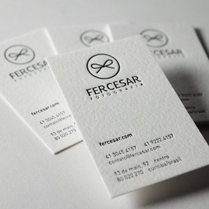 以下が含まれることがあります： 質感のあるオフホワイトの名刺の束。各カードには、FERCESAR FOTOGRAFIAという文字の上に、リボンのアイコンが入った黒い円形のロゴが印刷されています。ウェブサイト、電話番号、住所などの連絡先情報がその下に記載されています。