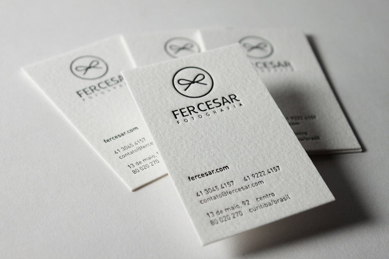 以下が含まれることがあります： 質感のあるオフホワイトの名刺の束。各カードには、FERCESAR FOTOGRAFIAという文字の上に、リボンのアイコンが入った黒い円形のロゴが印刷されています。ウェブサイト、電話番号、住所などの連絡先情報がその下に記載されています。