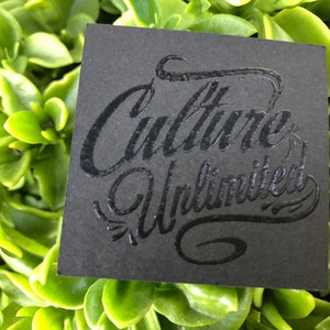 May include: A square black card featuring the embossed text Culture Unlimited in a stylized script font. The lettering is raised and glossy, contrasting with the matte texture of the card surface. The card is positioned against a background of green leaves.