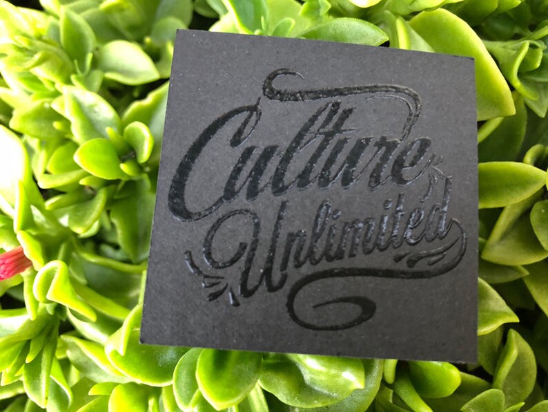 May include: A square black card featuring the embossed text Culture Unlimited in a stylized script font. The lettering is raised and glossy, contrasting with the matte texture of the card surface. The card is positioned against a background of green leaves.