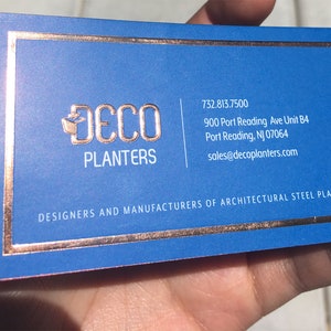 May include: A business card with a blue background and copper-colored text and border. The card features the logo "DECO PLANTERS" and contact information. The text "DESIGNERS AND MANUFACTURERS OF ARCHITECTURAL STEEL PLANTERS" is also visible.