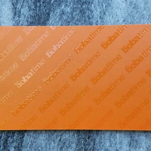 May include: A rectangular orange sheet featuring a repeating pattern of the word bobatime embossed across the surface in diagonal lines. The text is oriented in a consistent direction, creating a textured appearance on the flat orange material.