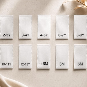 May include: A set of ten rectangular white fabric clothing labels arranged in two rows. Each label features black printed text indicating age sizes: 2-3Y, 3-4Y, 4-5Y, 6-7Y, 8-9Y, 10-11Y, 12-13Y, 0-6M, 3M, and 6M. The labels have a woven texture.