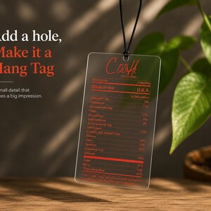 Puede incluir: Una etiqueta colgante rectangular de acr&iacute;lico transparente suspendida por un cord&oacute;n negro. La etiqueta presenta texto impreso en rojo que se asemeja a una etiqueta de informaci&oacute;n nutricional con la marca Coah Clothing y varios atributos.