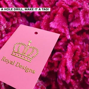 Puede incluir: Una etiqueta rectangular rosa con un icono de corona dorada y el texto Royal Designs en letras doradas. La etiqueta tiene un agujero preperforado en la parte superior central. El texto ADD A HOLE DRILL, MAKE IT A TAG! es visible arriba a la izquierda.