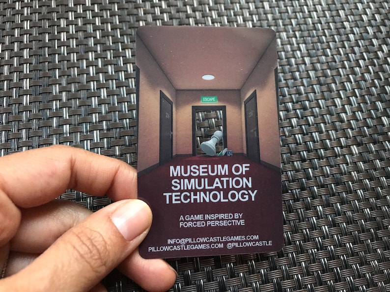 Puede incluir: Una tarjeta de juego rectangular con una ilustraci&oacute;n de un pasillo con una pieza de ajedrez blanca, el texto MUSEUM OF SIMULATION TECHNOLOGY, A GAME INSPIRED BY FORCED PERSPECTIVE, e informaci&oacute;n de contacto como INFO@PILLOWCASTLEGAMES.COM y PILLOWCASTLEGAMES.COM.
