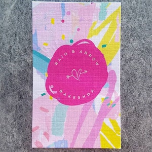 May include: A pink and white business card with a pink circle in the center. The circle has the text "RAIN & ARROW BAKESHOP" in white lettering with a white heart and arrow inside the circle.
