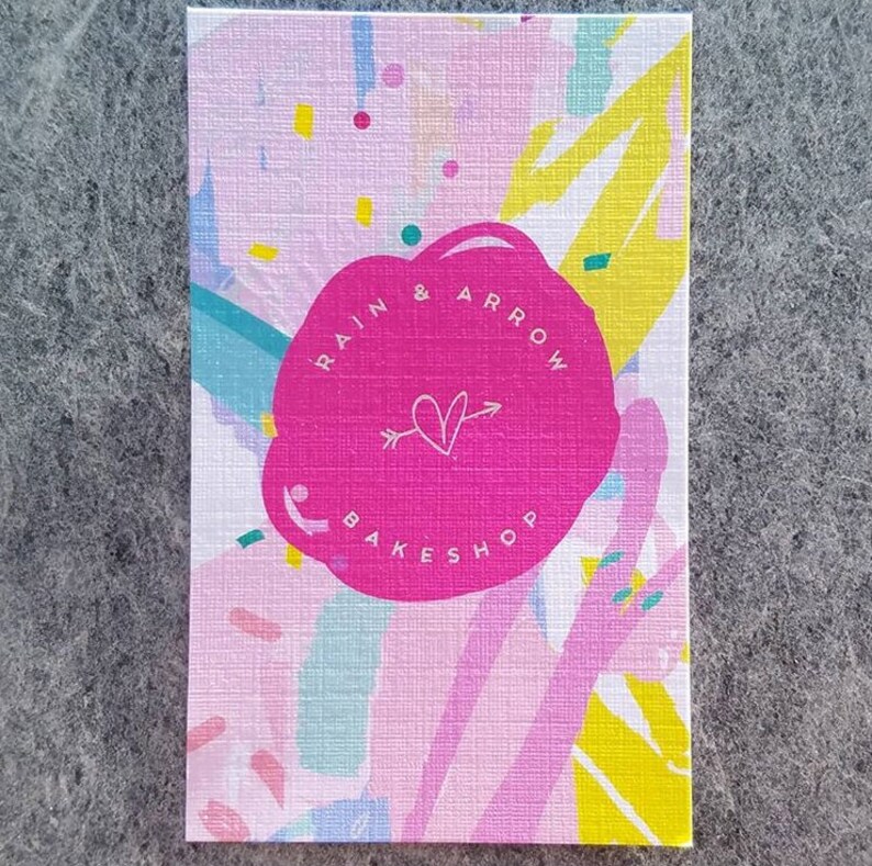 May include: A pink and white business card with a pink circle in the center. The circle has the text "RAIN & ARROW BAKESHOP" in white lettering with a white heart and arrow inside the circle.