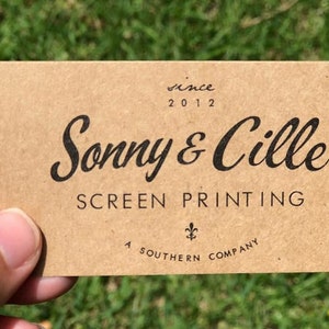 May include: A rectangular tan business card with black text that reads "since 2012", "Sonny & Cille", "SCREEN PRINTING", a small fleur-de-lis symbol, and "A SOUTHERN COMPANY" printed in a mix of cursive and sans-serif fonts on the front surface.