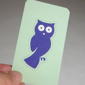 May include: A rectangular light green card featuring a dark purple owl silhouette in the center. The owl has two large white circular eyes with black pupils and a small white beak. A dashed line outlines the shape of the owl on the card.