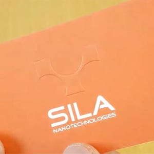 May include: A rectangular orange business card featuring a debossed logo at the top and the white printed text SILA NANOTECHNOLOGIES centered below. The card has a smooth matte finish and is held against a light-colored wooden surface background.