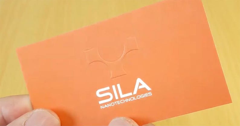 May include: A rectangular orange business card featuring a debossed logo at the top and the white printed text SILA NANOTECHNOLOGIES centered below. The card has a smooth matte finish and is held against a light-colored wooden surface background.