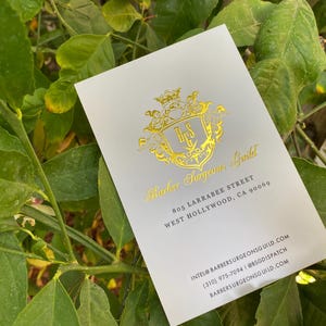 May include: A white rectangular business card featuring a gold foil crest logo with the letters BSG. The text reads Barber Surgeons Guild, 805 Larrabee Street, West Hollywood, CA 90069, intel@barbersurgeonsguild.com, (310) 975-7094, and barbersurgeonsguild.com.