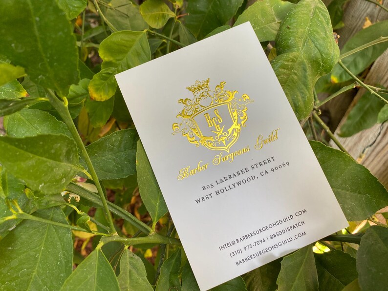 May include: A white rectangular business card featuring a gold foil crest logo with the letters BSG. The text reads Barber Surgeons Guild, 805 Larrabee Street, West Hollywood, CA 90069, intel@barbersurgeonsguild.com, (310) 975-7094, and barbersurgeonsguild.com.