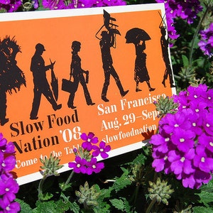 May include: A rectangular orange sign with black silhouettes of people carrying various items, featuring the text Slow Food Nation '08, San Francisco, Aug. 29-Sept. 1, and slowfoodnation.org, positioned among vibrant purple verbena flowers.