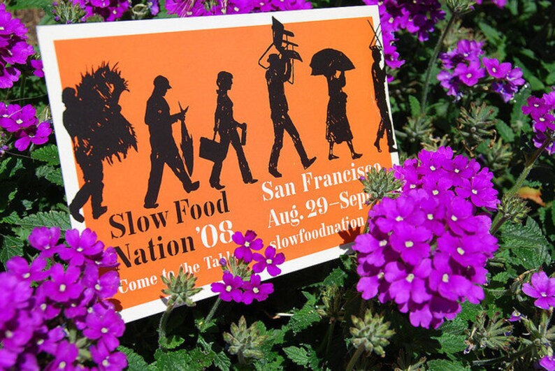 May include: A rectangular orange sign with black silhouettes of people carrying various items, featuring the text Slow Food Nation '08, San Francisco, Aug. 29-Sept. 1, and slowfoodnation.org, positioned among vibrant purple verbena flowers.