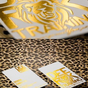 May include: Two rectangular white business cards featuring gold foil stamping. One card displays the text DEE ROCK TRAINING with a lion graphic, while the other includes the name DEREK CHRISTIE and additional contact information in gold text.
