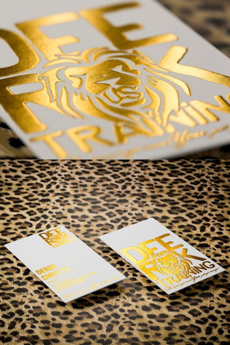 May include: Two rectangular white business cards featuring gold foil stamping. One card displays the text DEE ROCK TRAINING with a lion graphic, while the other includes the name DEREK CHRISTIE and additional contact information in gold text.