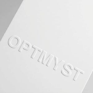 May include: A rectangular white card featuring the word OPTMYST embossed in capital letters across the center. The lettering is raised, creating a subtle three-dimensional effect against the smooth, matte white surface of the card stock.