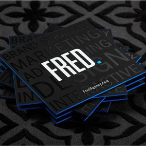 May include: A stack of square black business cards with blue edges. The top card features the white text FRED. and the website FredAgency.com, with additional black embossed text reading BRAND STRATEGY, MARKETING, and INTERACTIVE.