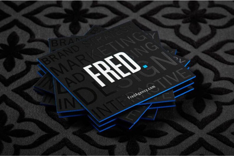 May include: A stack of square black business cards with blue edges. The top card features the white text FRED. and the website FredAgency.com, with additional black embossed text reading BRAND STRATEGY, MARKETING, and INTERACTIVE.