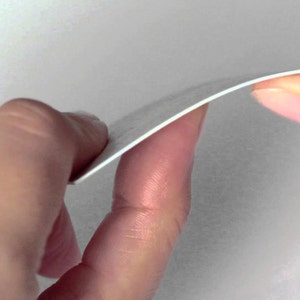 May include: A close-up of a white, slightly curved piece of paper being held between two hands. The paper appears to be thin and flexible, with a smooth surface. The edges are clean and well-defined.