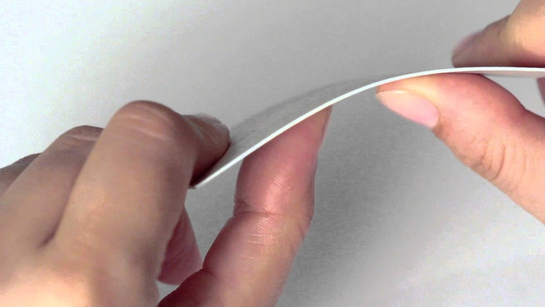 May include: A close-up of a white, slightly curved piece of paper being held between two hands. The paper appears to be thin and flexible, with a smooth surface. The edges are clean and well-defined.