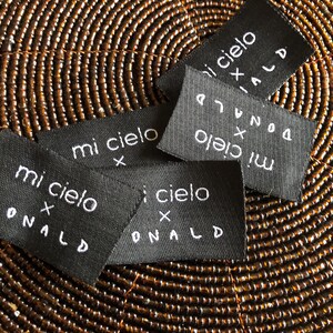 May include: A collection of rectangular black fabric labels featuring white embroidered text that reads mi cielo x DONALD. The labels are arranged in a scattered overlapping pattern, showcasing the consistent white lettering on the dark fabric.