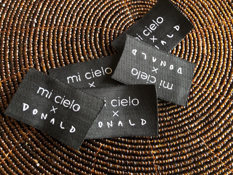 May include: A collection of rectangular black fabric labels featuring white embroidered text that reads mi cielo x DONALD. The labels are arranged in a scattered overlapping pattern, showcasing the consistent white lettering on the dark fabric.