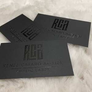 May include: Three rectangular black business cards featuring a glossy embossed logo with the letters RCA. Below the logo, the text reads RENEE CAFARO ATELIER and PUBLIC LABEL in a clean, sans-serif font. The cards have a matte finish and sharp edges.