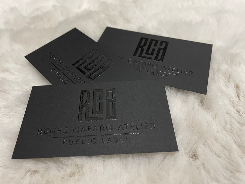 May include: Three rectangular black business cards featuring a glossy embossed logo with the letters RCA. Below the logo, the text reads RENEE CAFARO ATELIER and PUBLIC LABEL in a clean, sans-serif font. The cards have a matte finish and sharp edges.