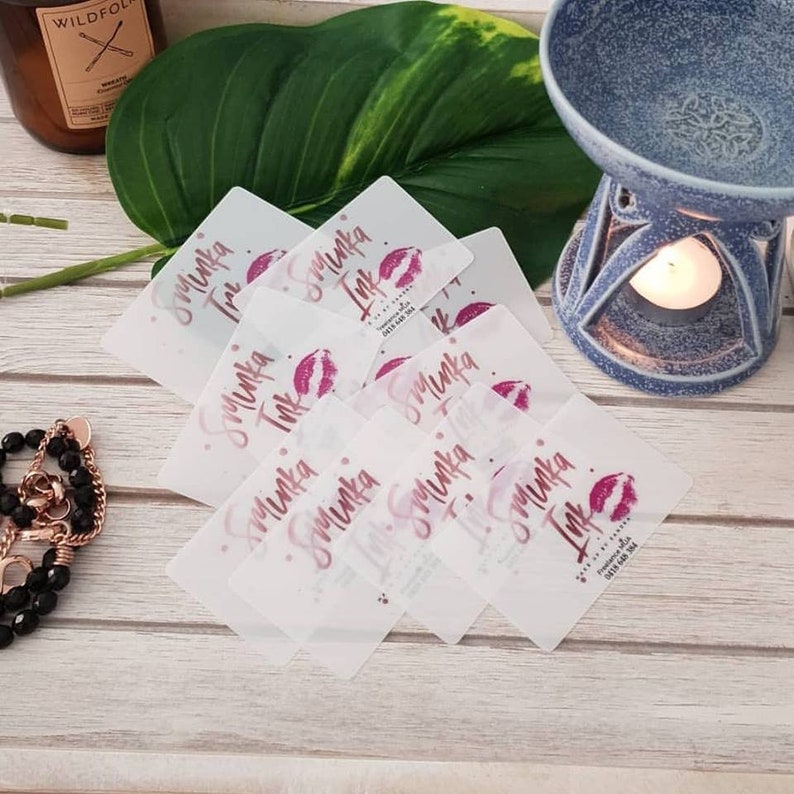May include: A stack of rectangular, semi-transparent business cards featuring the text Sminka Ink, a pink lip print, and contact details. The cards are arranged in a pile, showcasing a consistent design with pink lettering on a frosted surface.