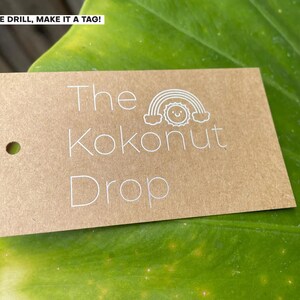 May include: A rectangular, light brown tag with the text "The Kokonut Drop" in white. A small white illustration of a rainbow and sun is above the word "Kokonut". The tag has a hole in the upper left corner. The tag is on a large green leaf.
