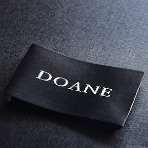 Puede incluir: Una etiqueta de ropa rectangular de tela negra con la palabra DOANE bordada en letras mayúsculas blancas. La etiqueta tiene una superficie lisa y ligeramente reflectante, colocada horizontalmente con un diseño sencillo y limpio.