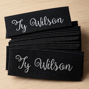 May include: A stack of rectangular black fabric labels featuring the name Ty Wilson embroidered in white cursive script. The labels are layered on top of each other, showcasing a consistent design with a clear, legible font on a dark background.