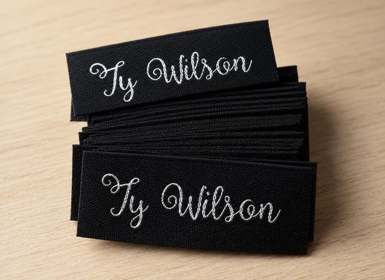 May include: A stack of rectangular black fabric labels featuring the name Ty Wilson embroidered in white cursive script. The labels are layered on top of each other, showcasing a consistent design with a clear, legible font on a dark background.
