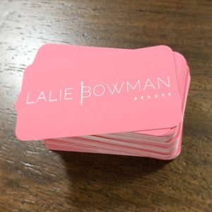 K&ouml;nnte beinhalten: Ein Stapel rosa Visitenkarten mit dem Text "Lalie Bowman Beauty" in wei&szlig;er Schrift.